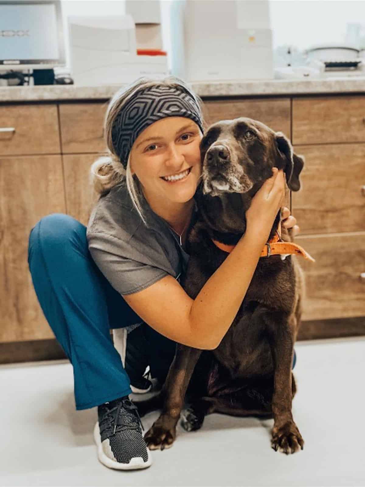 A smiling veterinarian in scrubs and a headband kneels on the floor inside a veterinary clinic, hugging a brown dog with a gray muzzle and orange collar. The clinic background includes cabinets and equipment.