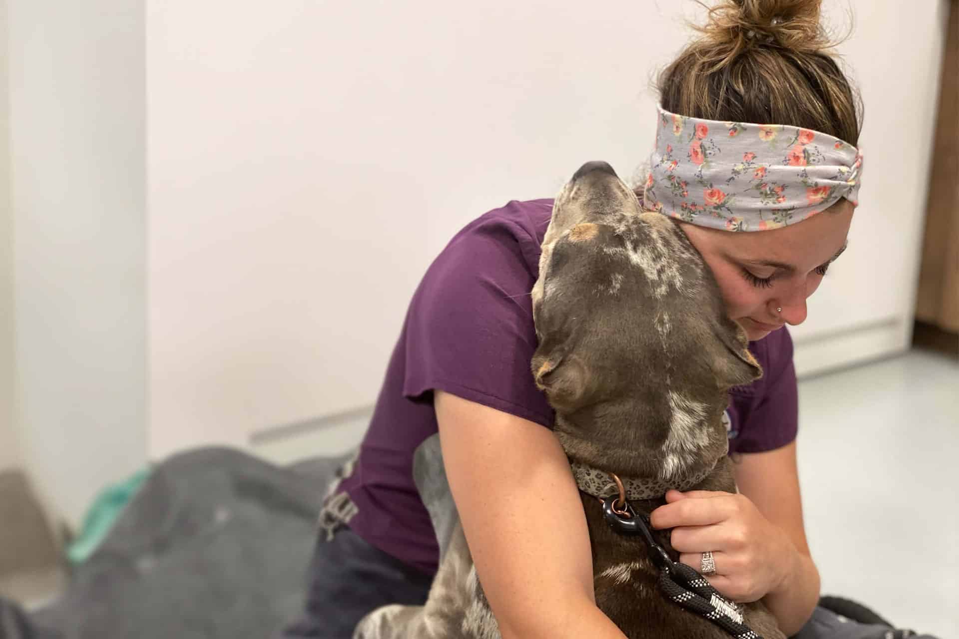 A woman in a purple shirt and floral headband hugs a gray and white dog. The woman’s eyes are closed, and the dog’s head rests on her shoulder. They are seated on a gray surface with a white wall in the background, creating a warm and affectionate moment, as if they just had a comforting visit to the vet.