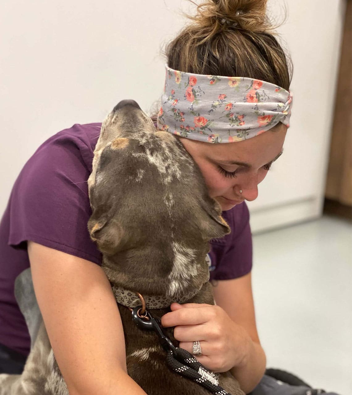 A woman wearing a floral headband embraces a gray and white dog with her eyes closed, like a relieved owner after a vet visit. The dog leans into her affectionately, its leash held in the woman's hand against the neutral indoor backdrop.