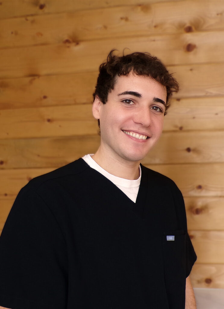 Jake A young man with short curly hair, wearing a black scrub top over a white shirt, smiles while standing in front of a wooden wall.