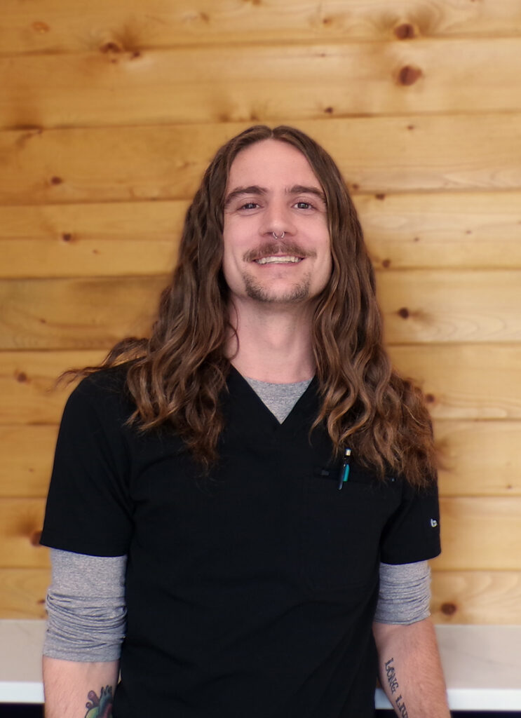 Josh A smiling person with long, wavy brown hair wearing a black scrub top over a gray long-sleeve shirt stands in front of a wooden wall.