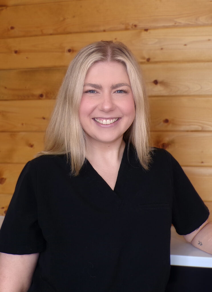Kathleen A smiling woman with straight blonde hair wearing a black scrub top stands in front of a wooden wall.