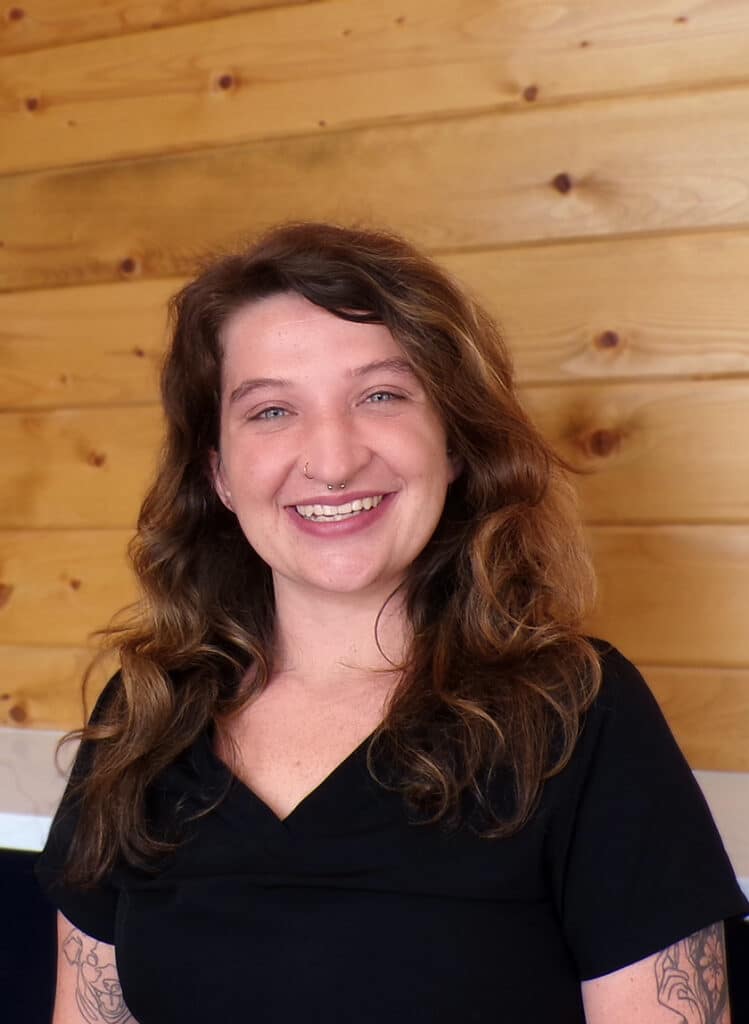 A smiling woman with wavy brown hair, wearing a black top, stands in front of a wooden wall. She has tattoos on her arm and a nose ring.