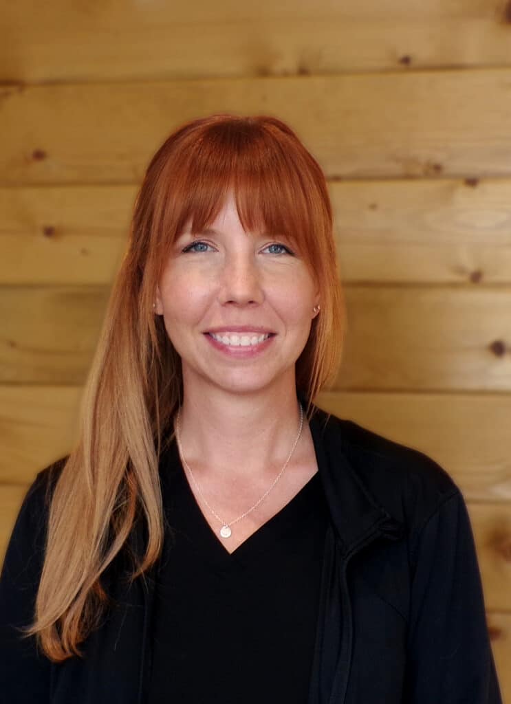 A woman with straight, long auburn hair and bangs smiles at the camera. She is wearing a black top and a necklace, standing in front of a wooden wall with horizontal panels.