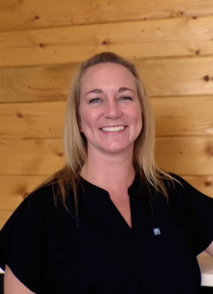 A woman with blonde hair wearing a black top smiles in front of a wooden wall background.