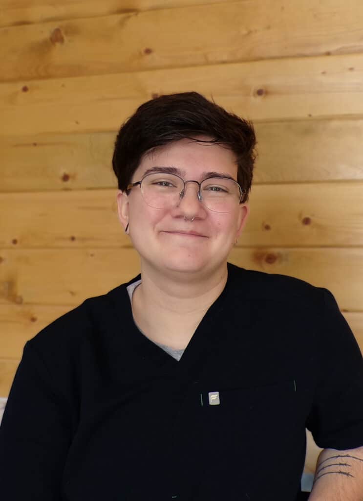 A person with short dark hair, glasses, and a nose ring smiles softly. They are wearing a black shirt, sitting in front of a wooden wall with horizontal panels.