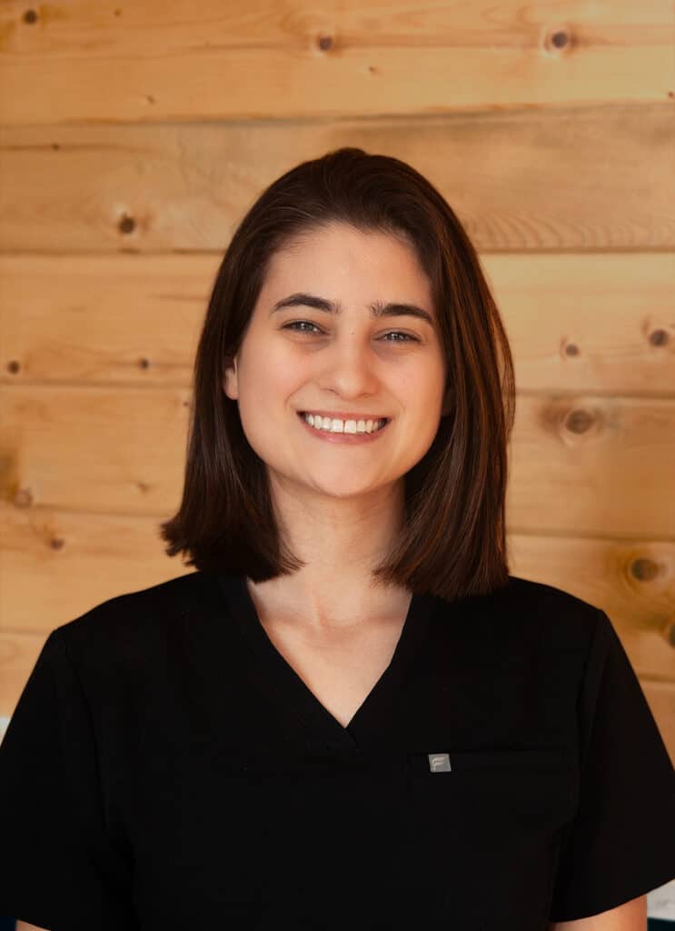 A woman with straight, shoulder-length brown hair smiles at the camera. She is wearing a black scrub top and stands in front of a wooden wall with a natural finish.