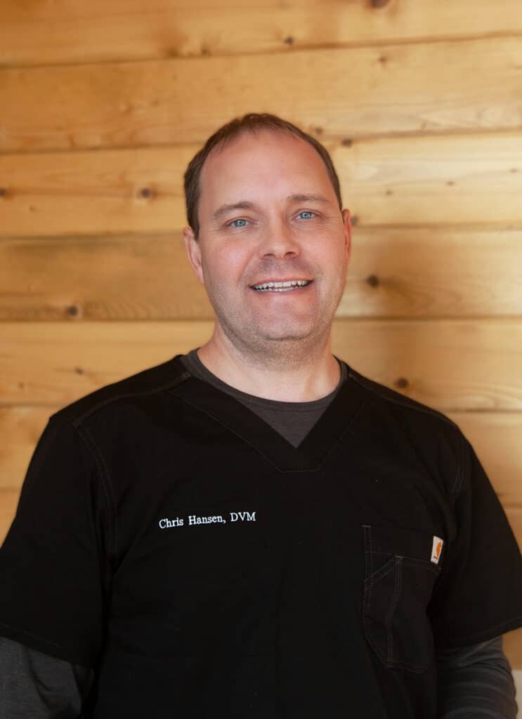 A man wearing a black medical scrub top with the embroidered name "Chris Hansen, DVM" smiles in front of a wooden wall background.