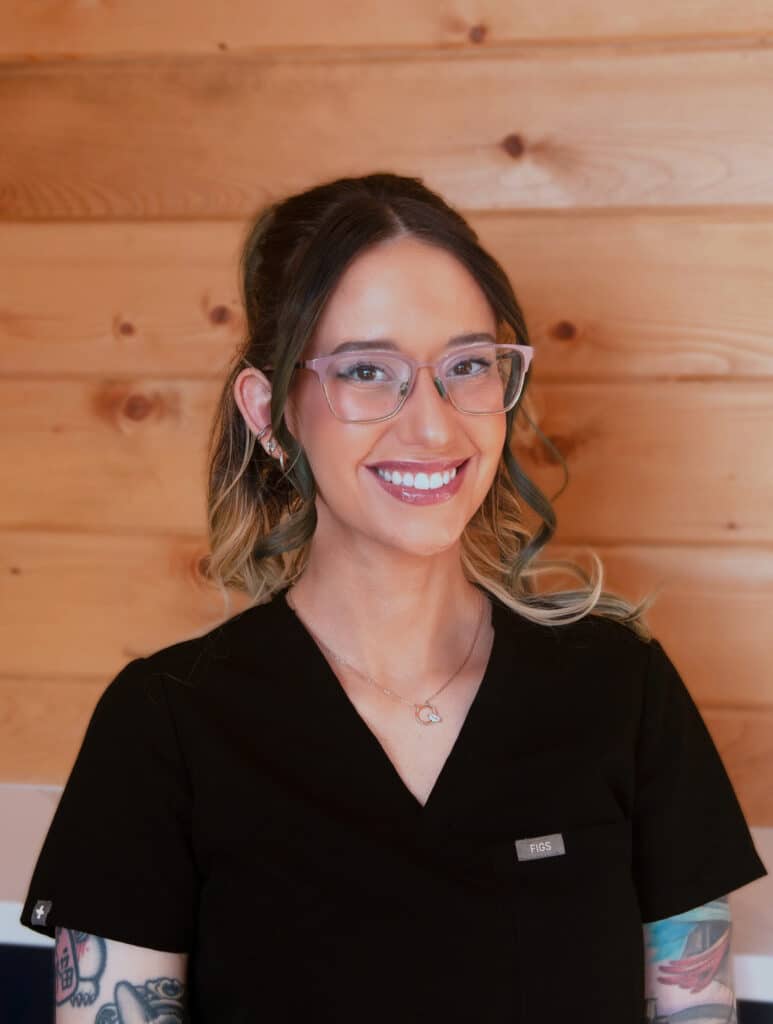 A woman with glasses and tattoos, wearing black scrubs and a necklace, smiles while standing in front of a wooden wall.