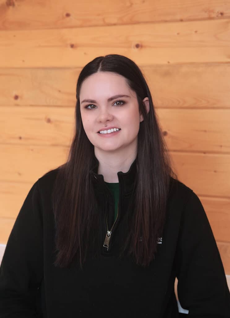 A woman with long dark hair, wearing a black zip-up jacket, smiles while sitting in front of a light-colored wooden wall.
