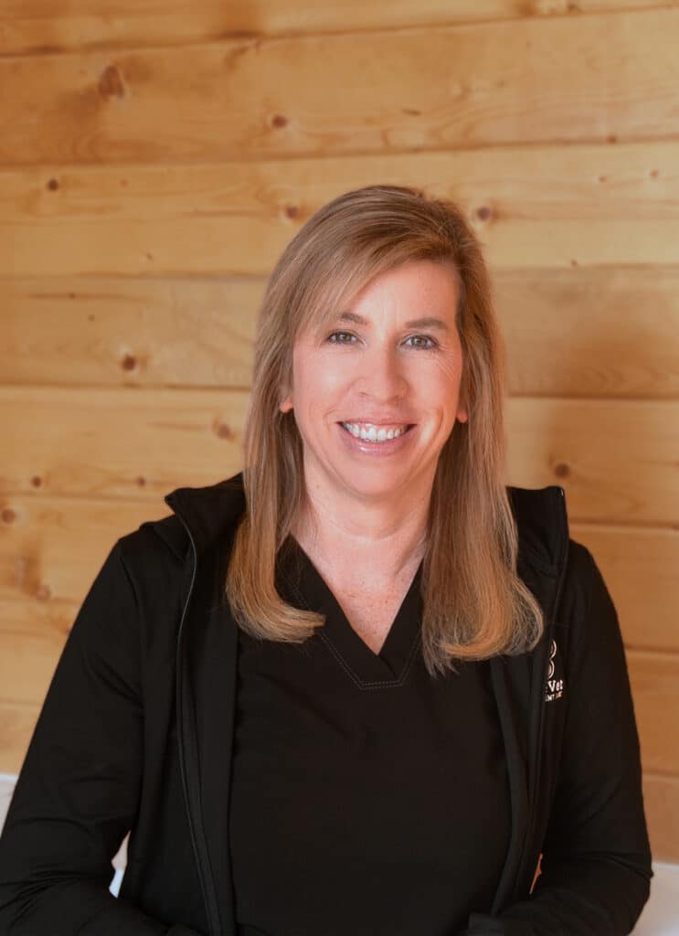 A woman with long blonde hair wearing a black shirt and jacket smiles while seated in front of a wooden wall.