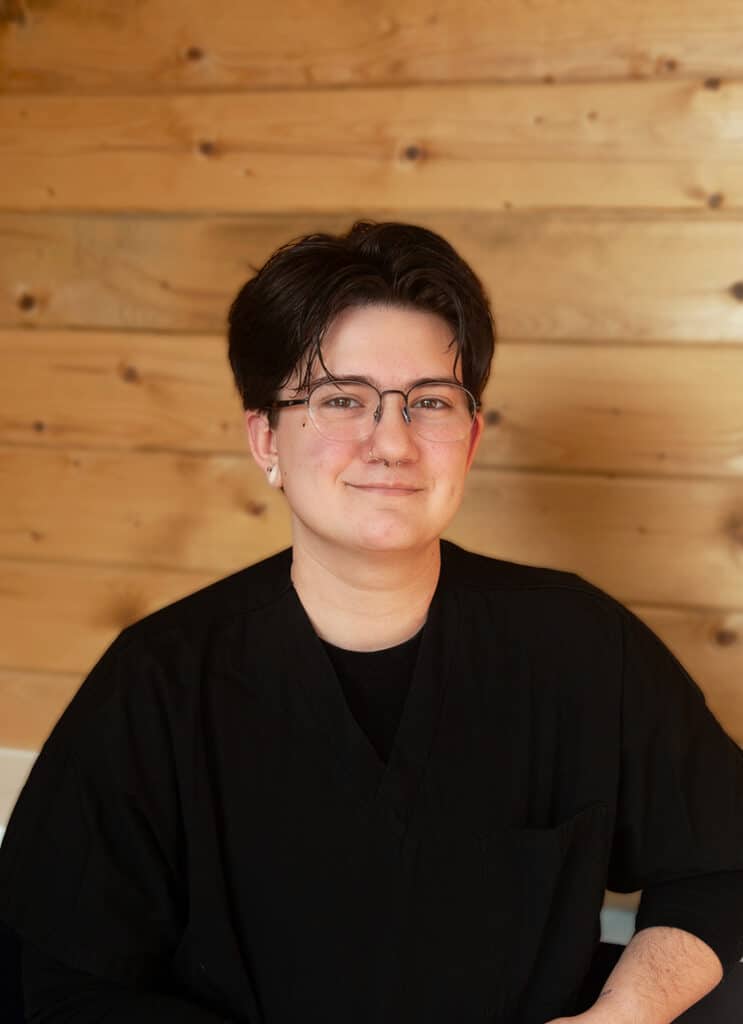 A person with short dark hair, glasses, and an earring is smiling softly. They are wearing a black top and are seated against a background of light wooden planks.