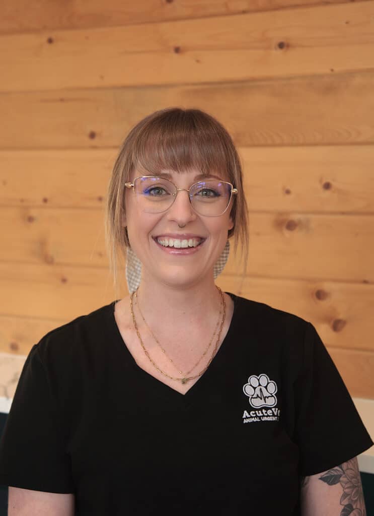 A smiling person with glasses and bangs, wearing a black scrub top with "AcuteVet Animal Urgent Care" and a paw print logo, stands in front of a wooden wall.