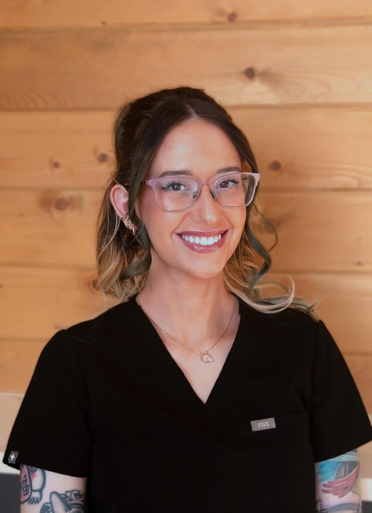 A smiling woman with glasses and tattoos, wearing a black shirt with a name badge, stands in front of a wooden wall background.