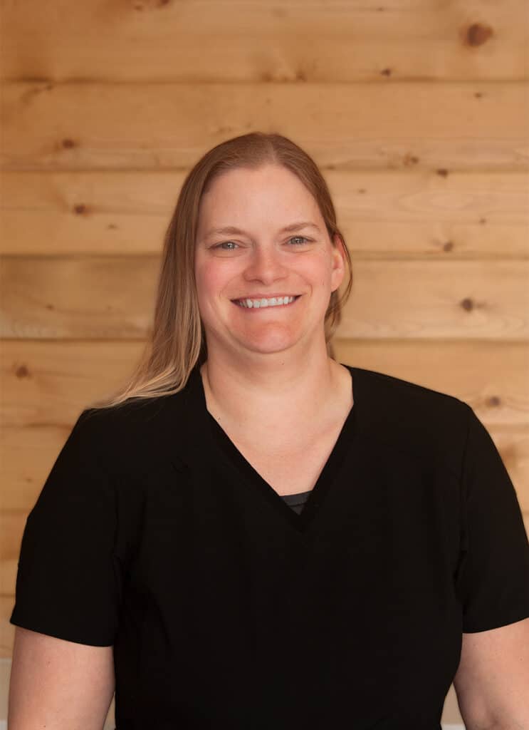 A woman with straight, light brown hair wearing a black shirt smiles at the camera while standing in front of a wooden, light-colored plank wall.