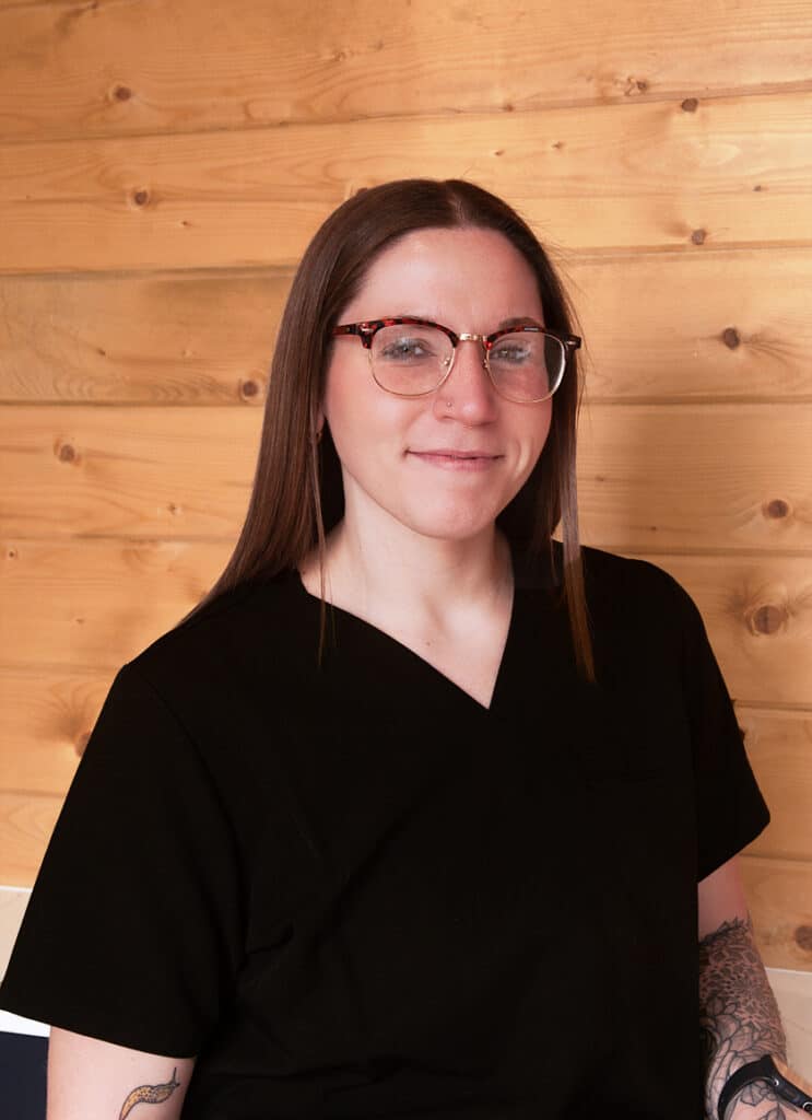 A person with long brown hair, glasses, and a black scrub top is smiling and sitting in front of a wooden wall. A tattoo is partially visible on their right arm.