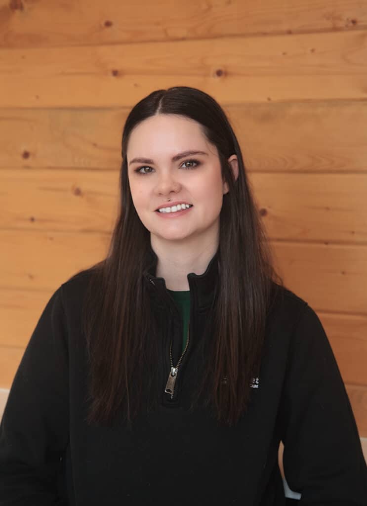 A young woman with long brown hair, wearing a black zip-up jacket, smiles while sitting in front of a wooden wall background.