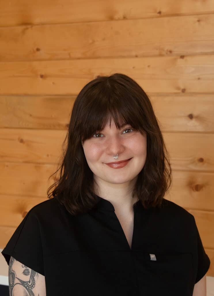 A person with shoulder-length dark brown hair, a nose ring, and a black top smiles in front of a wooden wall. Part of a tattoo is visible on their left arm.